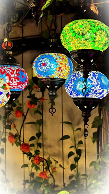The image shows a collection of colorful mosaic lamps hanging from the ceiling in an interior space with greenery and flowers visible through what app
