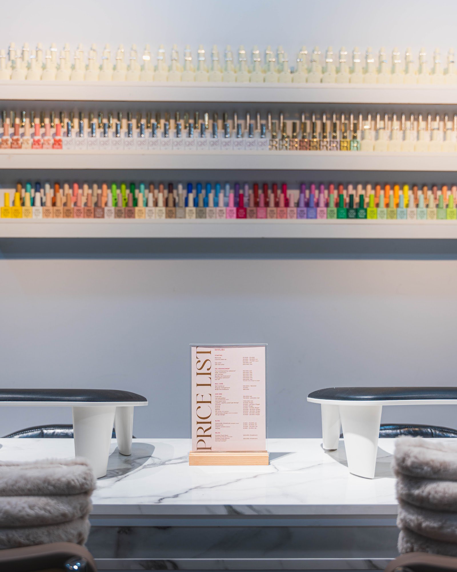 The image shows a neatly organized nail salon counter displaying an extensive array of colorful nail polish bottles on shelves above and below a price