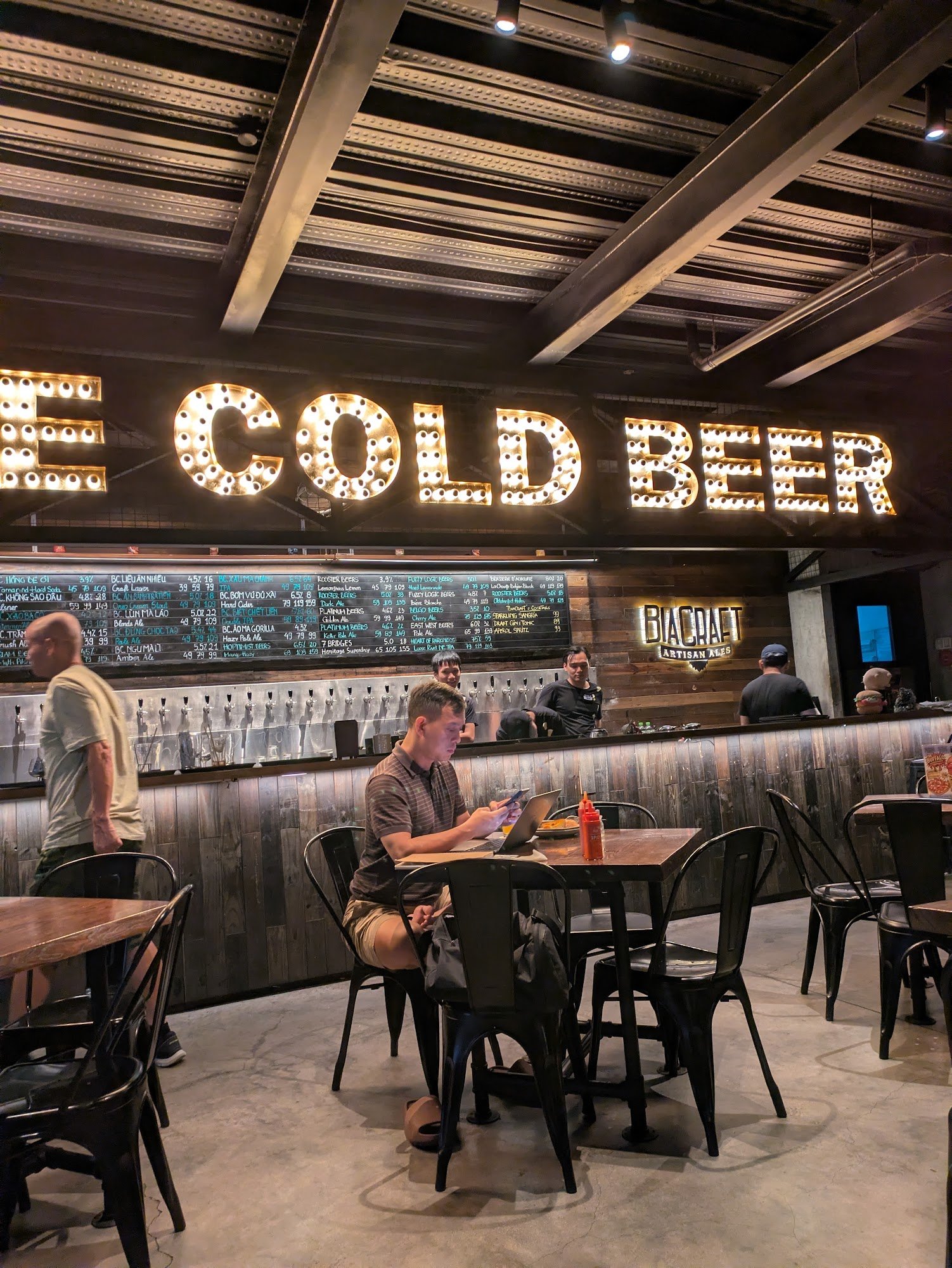 The image depicts the interior of a craft beer bar named 'BIACRAFT ARTISAN ALES' located in Thao Dien, Saigon. A prominent illuminated sign reading 'C