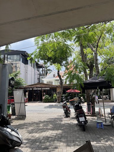 The image depicts an outdoor plaza in Thao Dien, Saigon. It features a paved area with several motorbikes parked, a building with a visible 'Cafe' sig