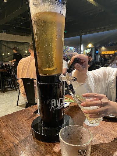 A close-up of a beer being poured from a tall glass into a smaller cup using a pump system attached to the larger glass, which is branded with 'BIACO'