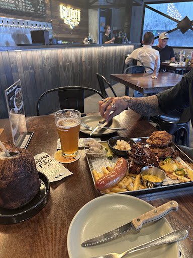 A restaurant interior in Thao Dien, Saigon featuring a wooden table with a large beer glass, a plate with a knife and fork, a tray of food including a