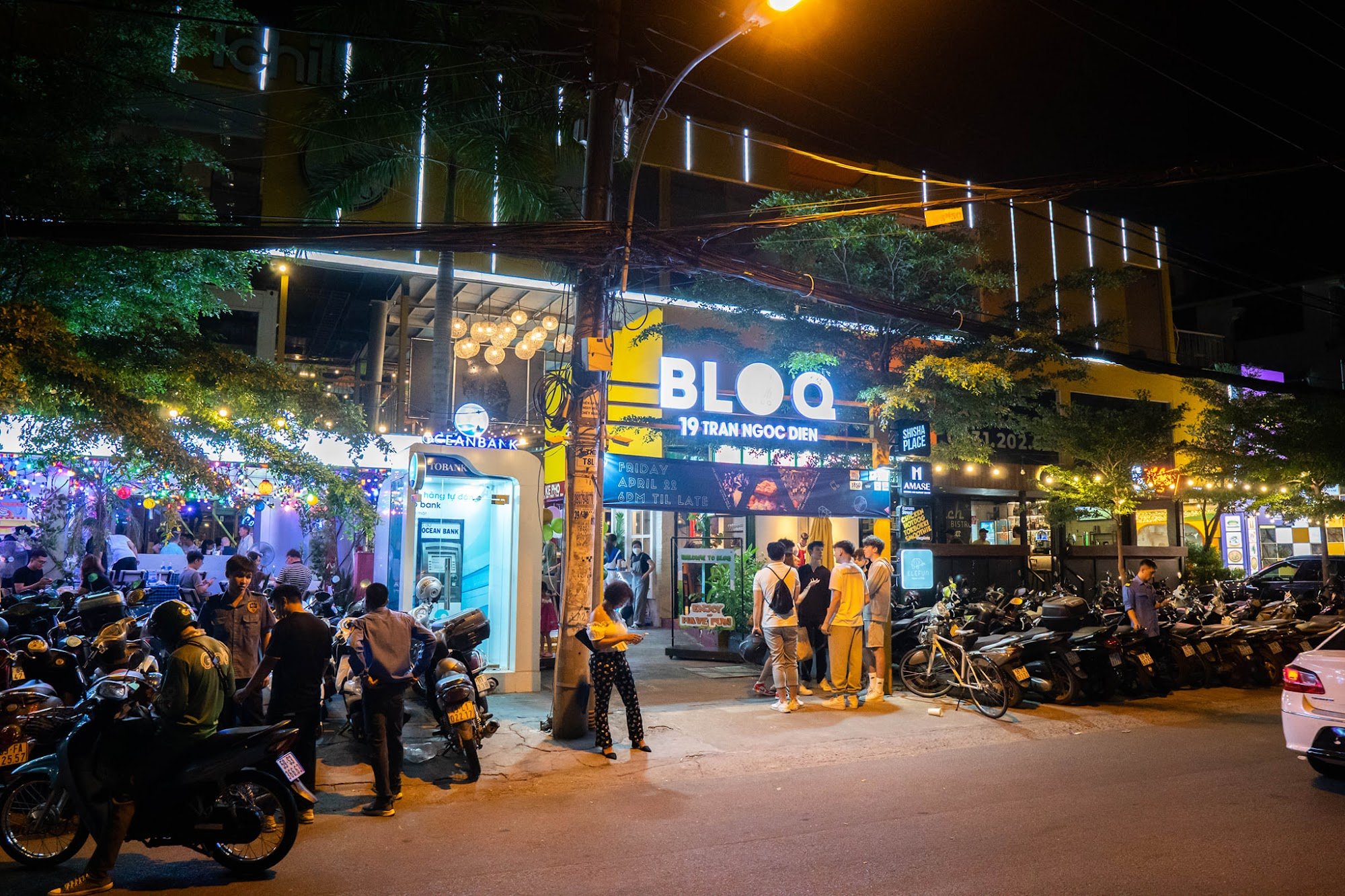 The image captures a bustling night scene outside an establishment named 'BLOQ' on Tran Ngoc Dien street in Thao Dien, Saigon. The area is illuminated