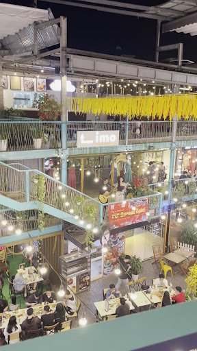 The image shows an interior view of a multi-level food court or market hall at night time with various dining areas on different levels, decorated wit