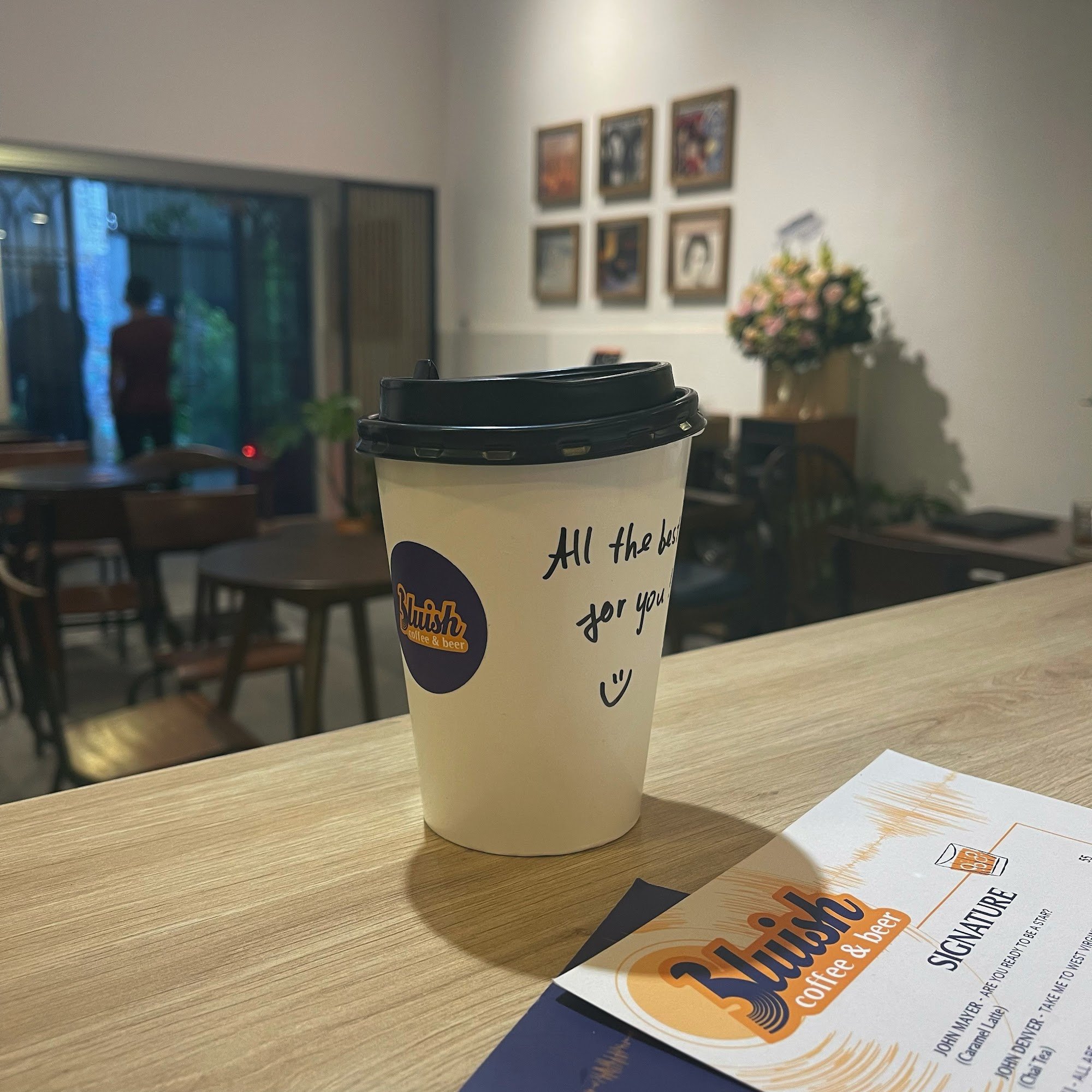 A disposable coffee cup from 'Buish Coffee & Beer' is placed on a wooden table in an indoor setting with ambient lighting and framed pictures on the w