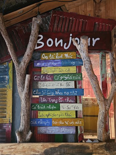 The image shows a colorful signboard displaying various phrases in different languages that say 'Bonjour'. The background features rustic wooden eleme
