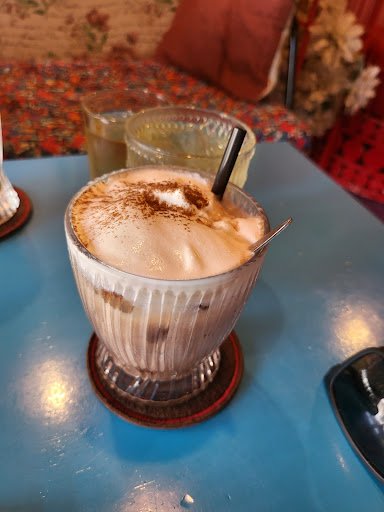 A frothy beverage topped with a dusting of cocoa powder and garnished with a long metal straw sits in a glass on a coaster, placed on a blue table.