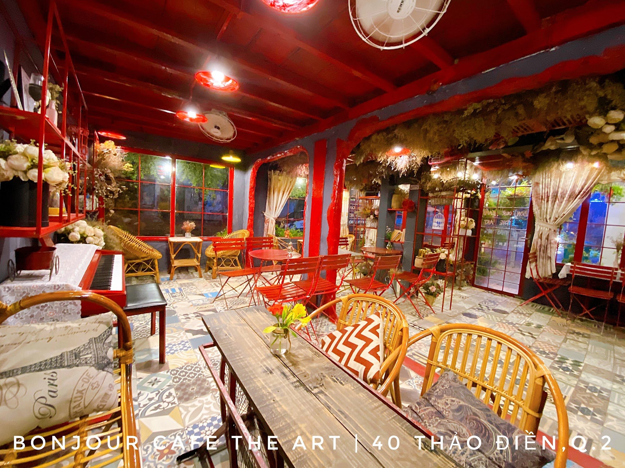 The image depicts the interior of Bonjour Cafe The Art, located at 40 Thao Dien, Q.2, Saigon. The space features vibrant red and blue walls with red m