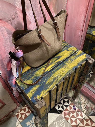 A beige handbag is placed on a colorful wooden crate that sits atop an ornate tiled floor with geometric patterns and contrasting colors. The backgrou
