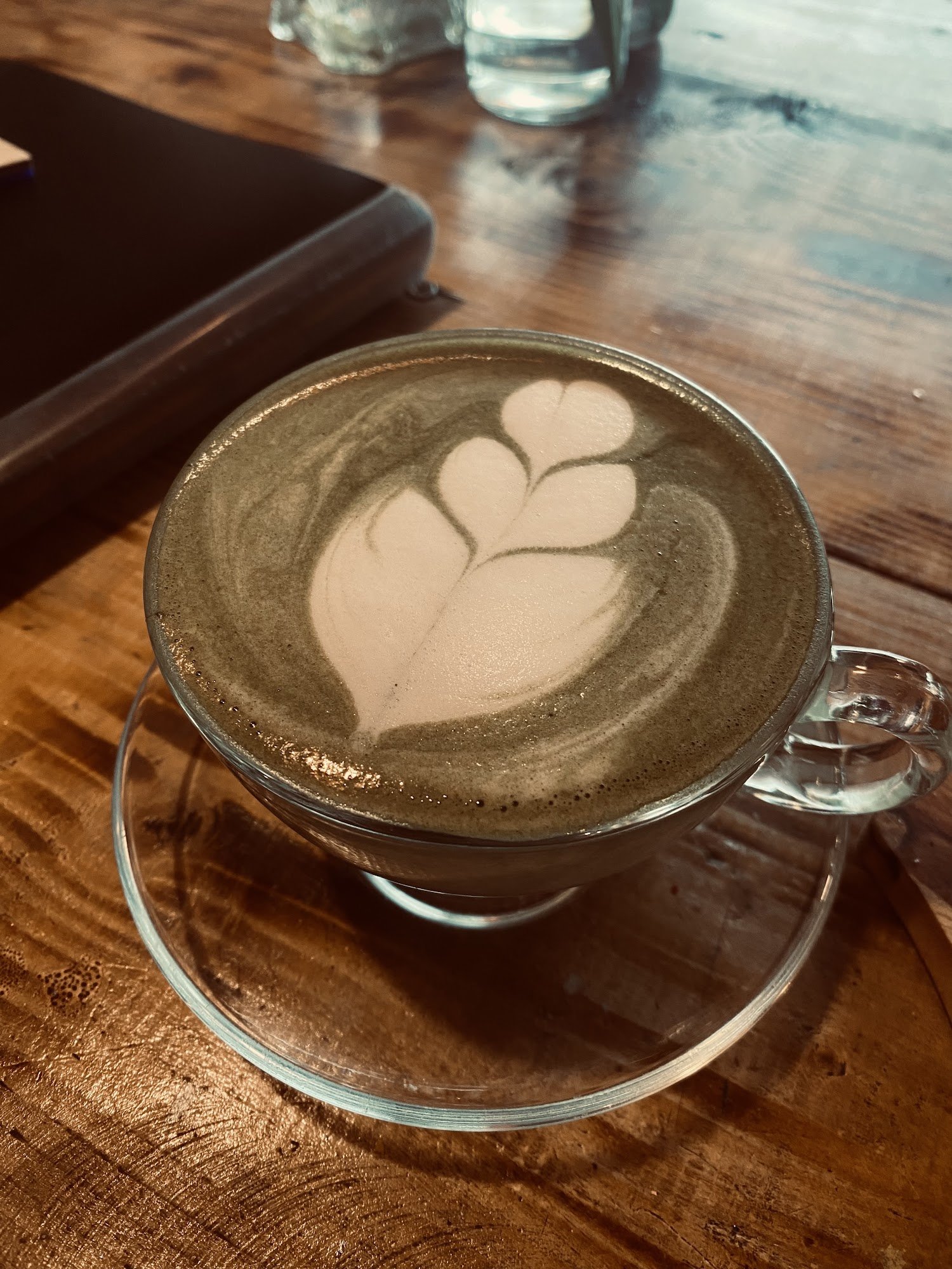 A close-up of a latte art on top of espresso in a clear glass cup with a handle and saucer placed on a wooden table, suggesting it might be from Thao 