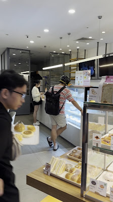 The image shows an indoor setting of a bakery or café where customers are browsing and purchasing items from the display case.