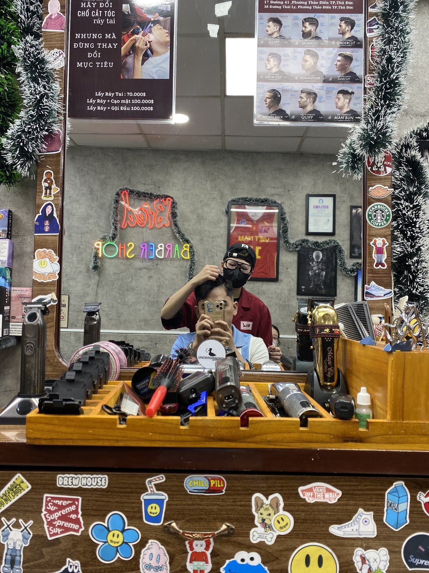 The image depicts the interior of a barber shop in Thao Dien, Saigon. A barber in a red shirt and black cap is styling a client's hair while taking a 