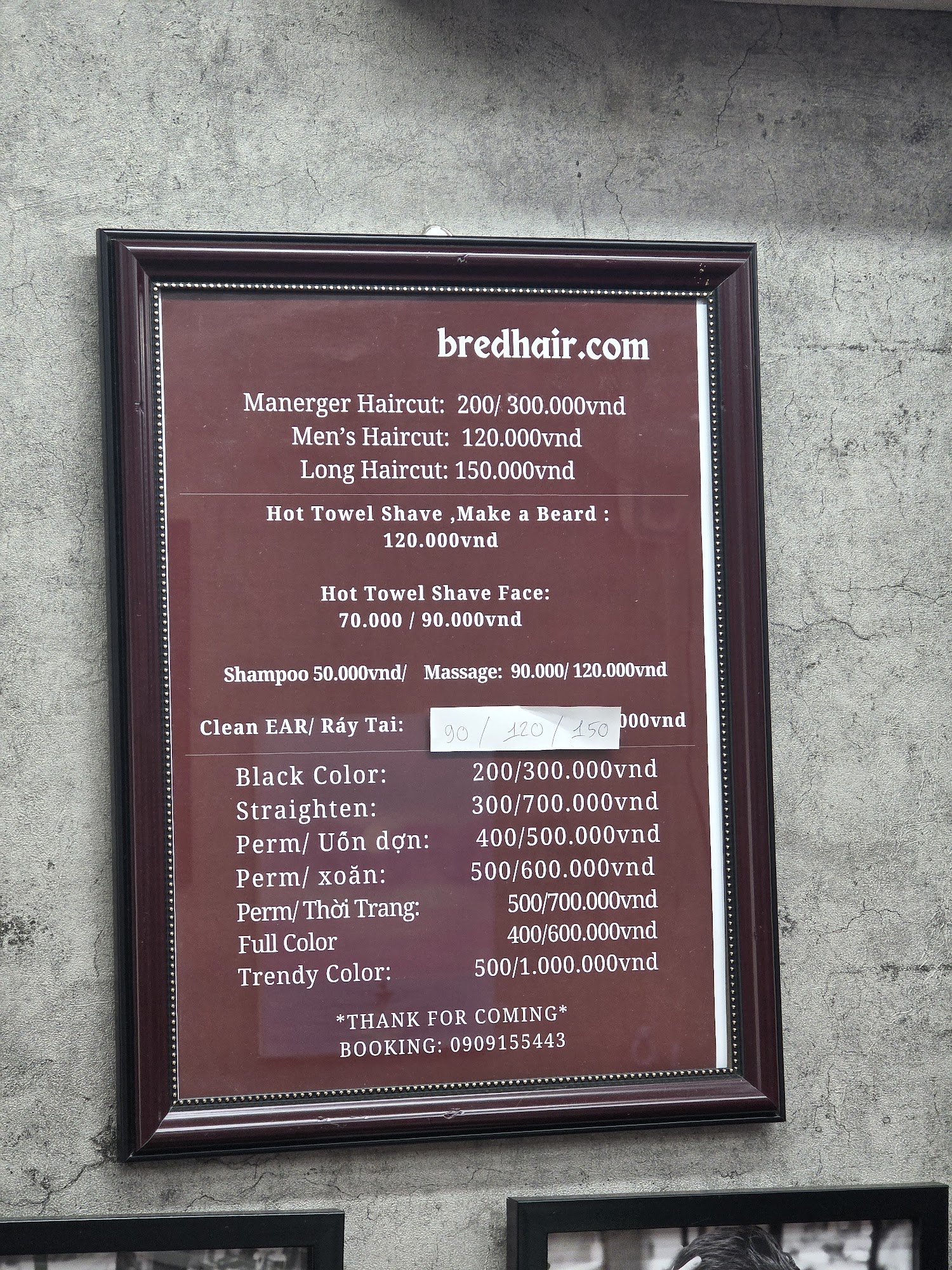 A framed menu board displaying hair salon services and prices in Vietnamese Dong (VND). The background is a textured concrete wall, suggesting an urba