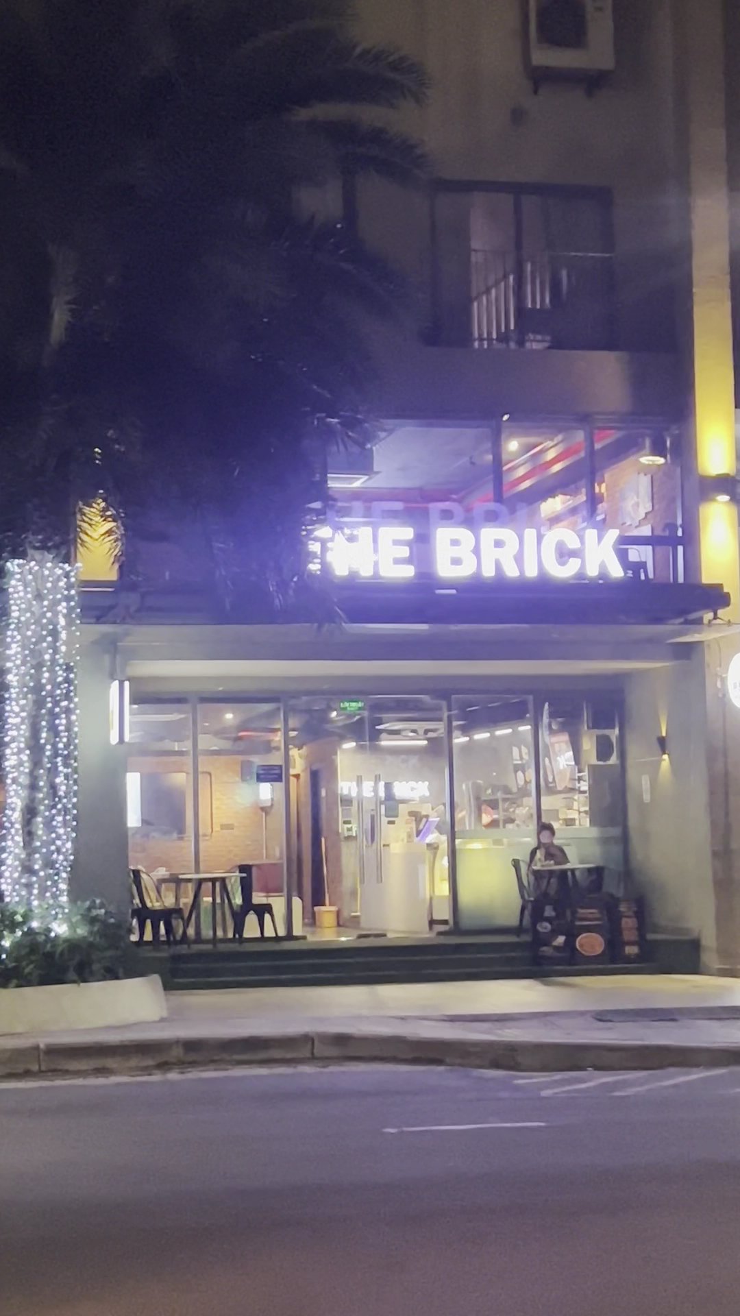 The image shows an exterior view of a restaurant named 'THE BRICK' during nighttime. The establishment is illuminated with bright lights, including ne