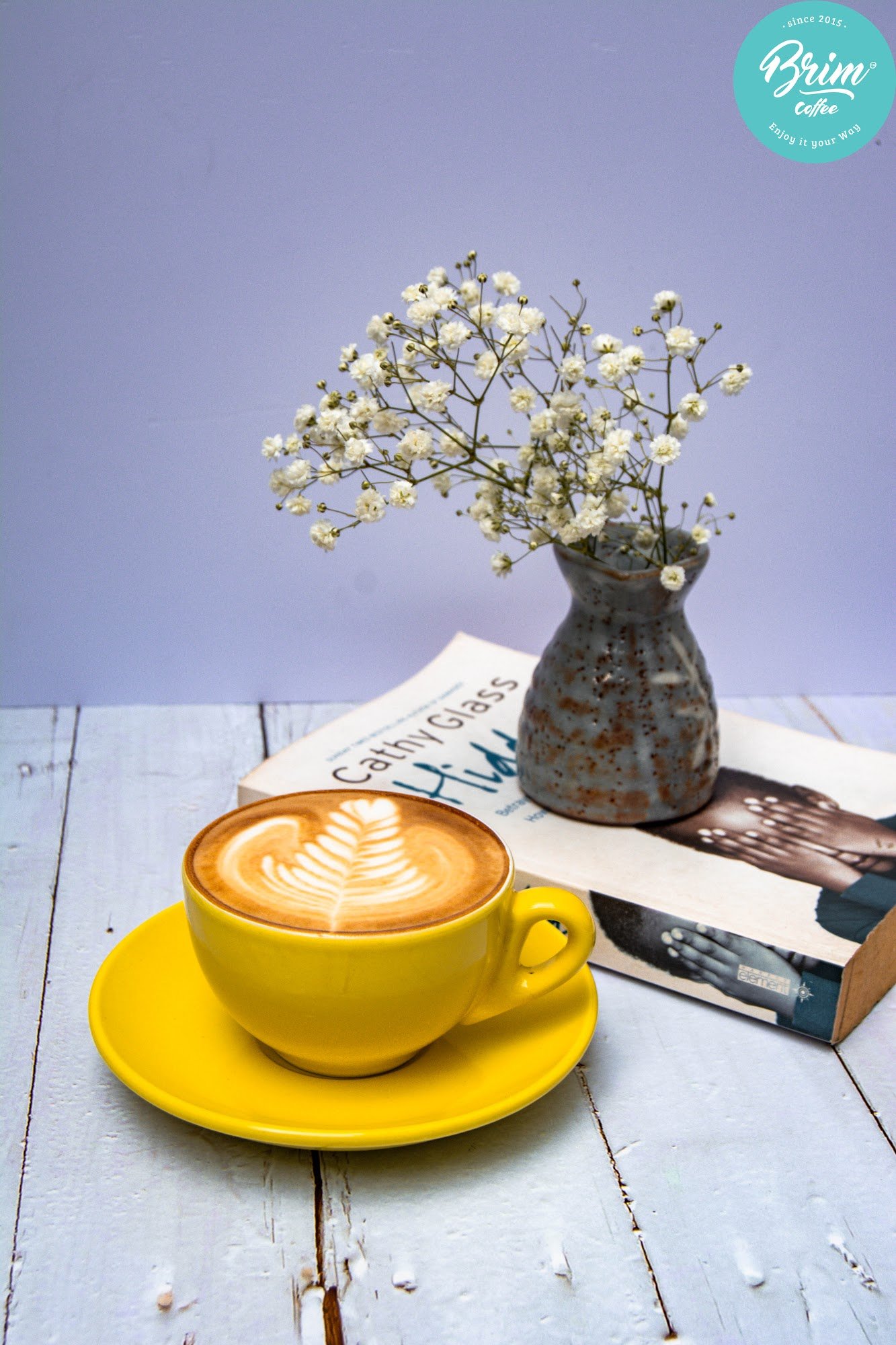 This image depicts a cozy interior setting featuring a cup of latte art on a yellow saucer with a matching plate, placed next to an open book titled '