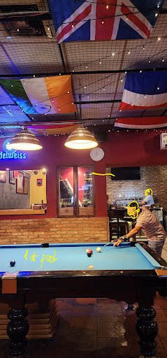 The image depicts an indoor pool hall featuring a blue billiards table in the foreground with several balls scattered across it and one player activel