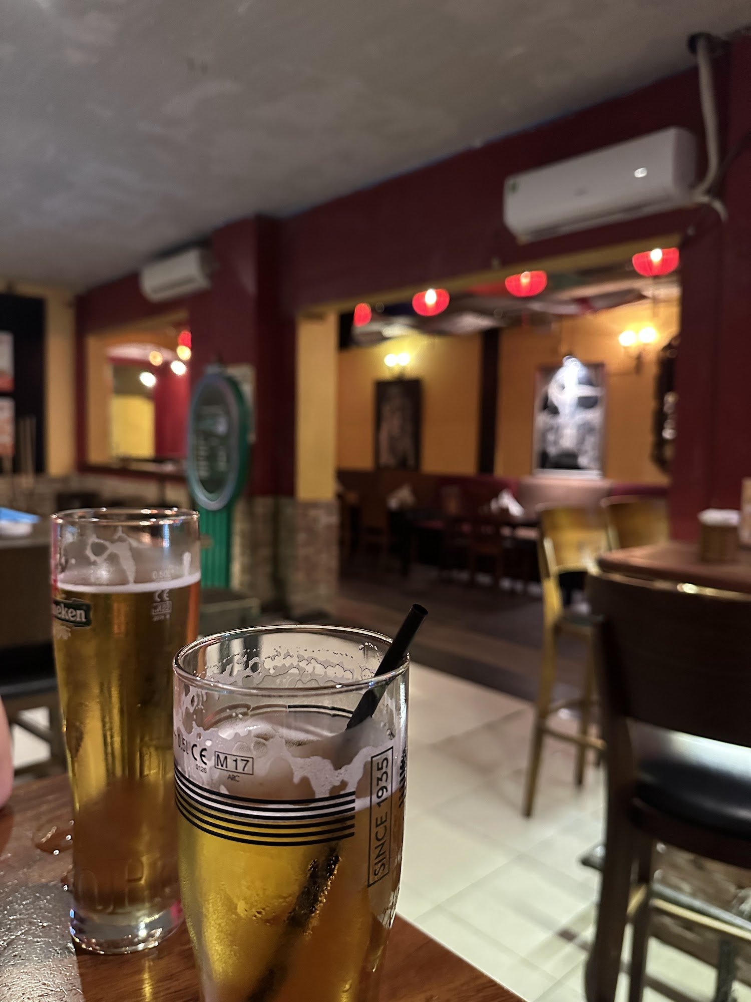 Two glasses of beer on a wooden table in a bar/restaurant interior. One glass is a tall Heineken beer glass, the other is a shorter glass with "SINCE