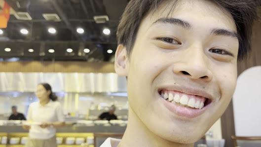 A close-up of a person in a restaurant setting, with background showing a dining area featuring tables, chairs, staff (a woman in white uniform), and