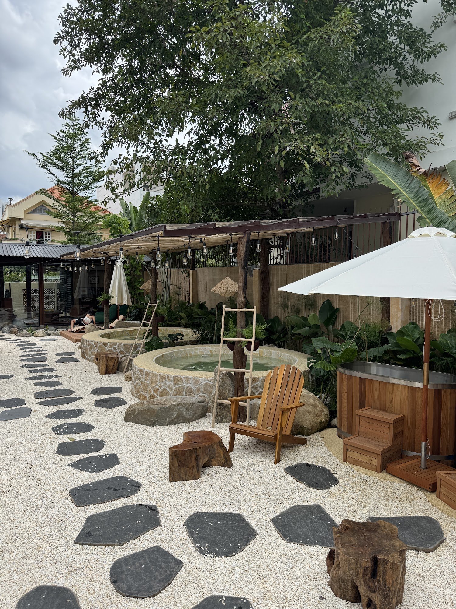 The image depicts an outdoor area of a venue in Thao Dien, Saigon. It features a hot tub surrounded by stepping stones and various seating options inc