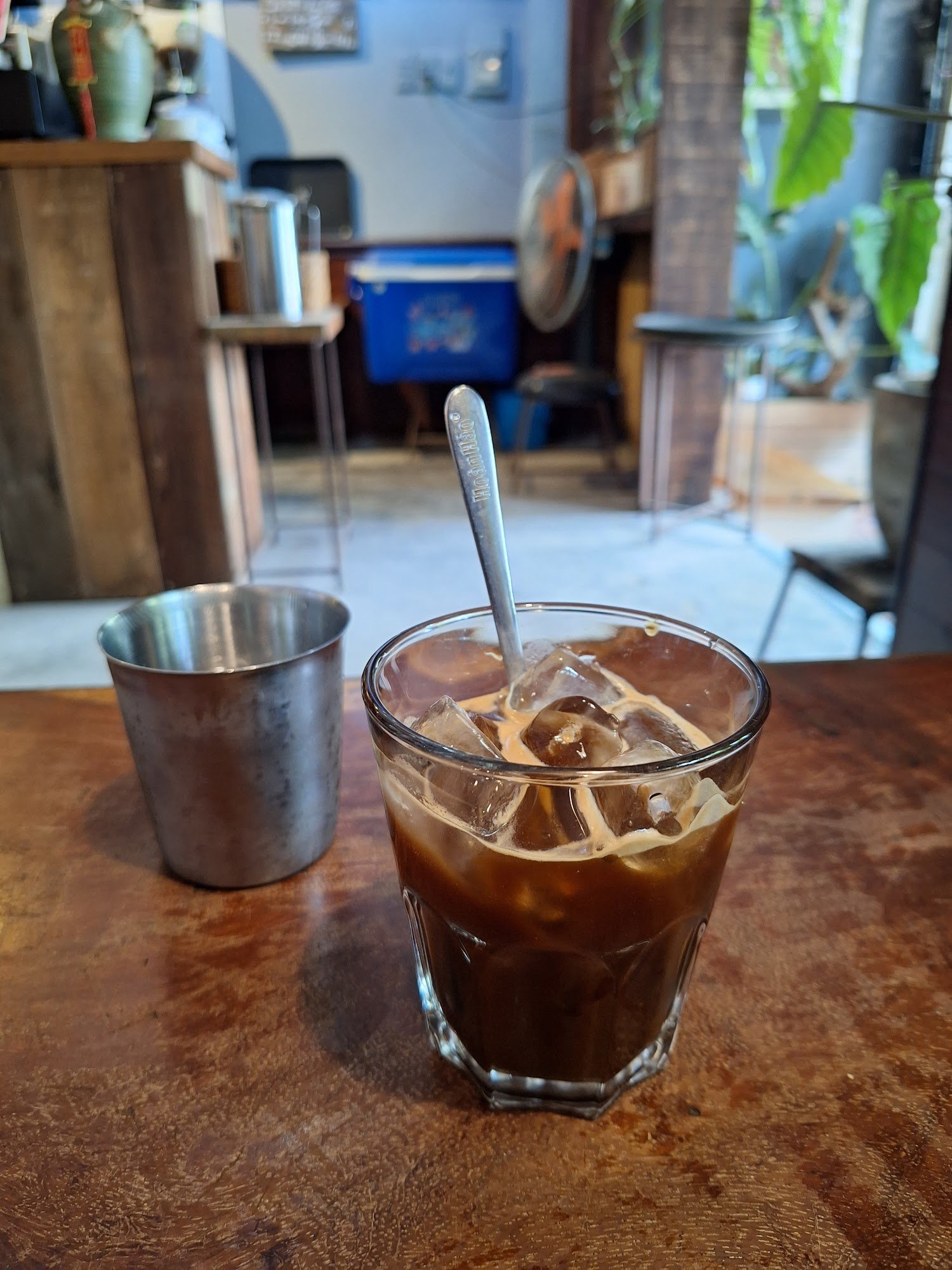A glass of cold coffee beverage on a wooden table in an indoor setting that appears to be a cafe or restaurant with various items and decor visible in