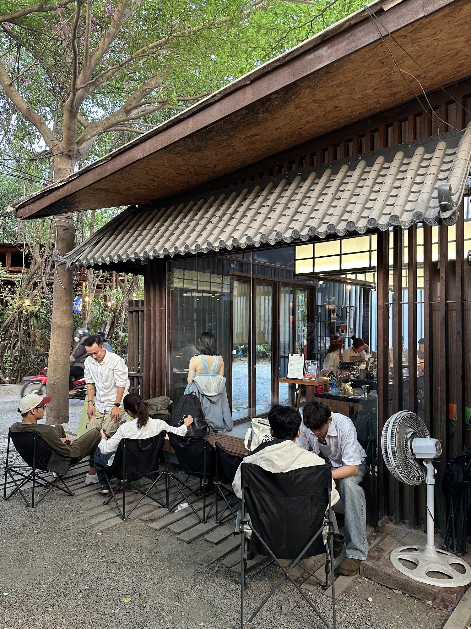 The image depicts an outdoor seating area of a café or restaurant in Thao Dien, Saigon. Several patrons are seated on folding chairs outside the estab