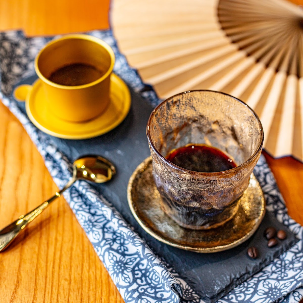 The image depicts a cozy corner of an establishment featuring two cups on a decorative tray with a fan and coffee beans beside them, suggesting a rela