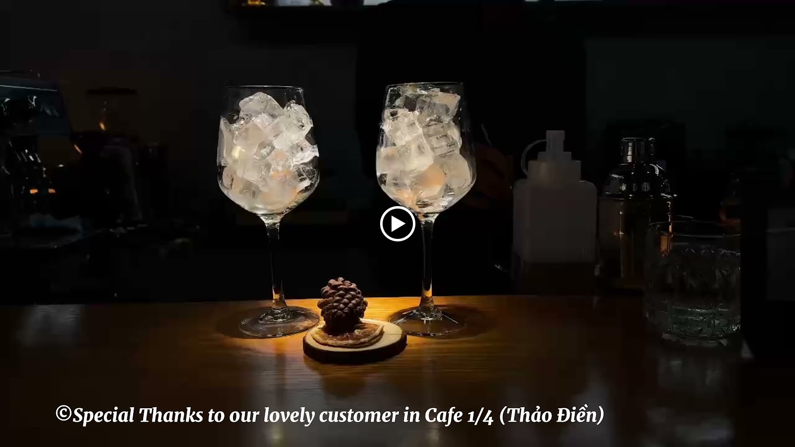 The image shows a dimly lit bar setting with two glasses filled with ice and what appears to be whiskey on the rocks, placed centrally on a wooden sur