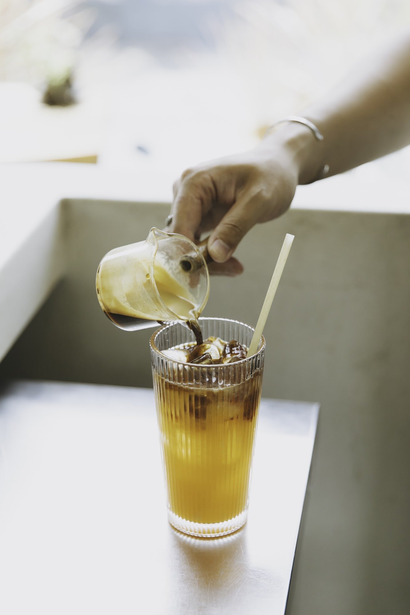 A hand is pouring a yellowish liquid from a small jug into a glass containing an already prepared drink with ice and chocolate syrup, which has a stra