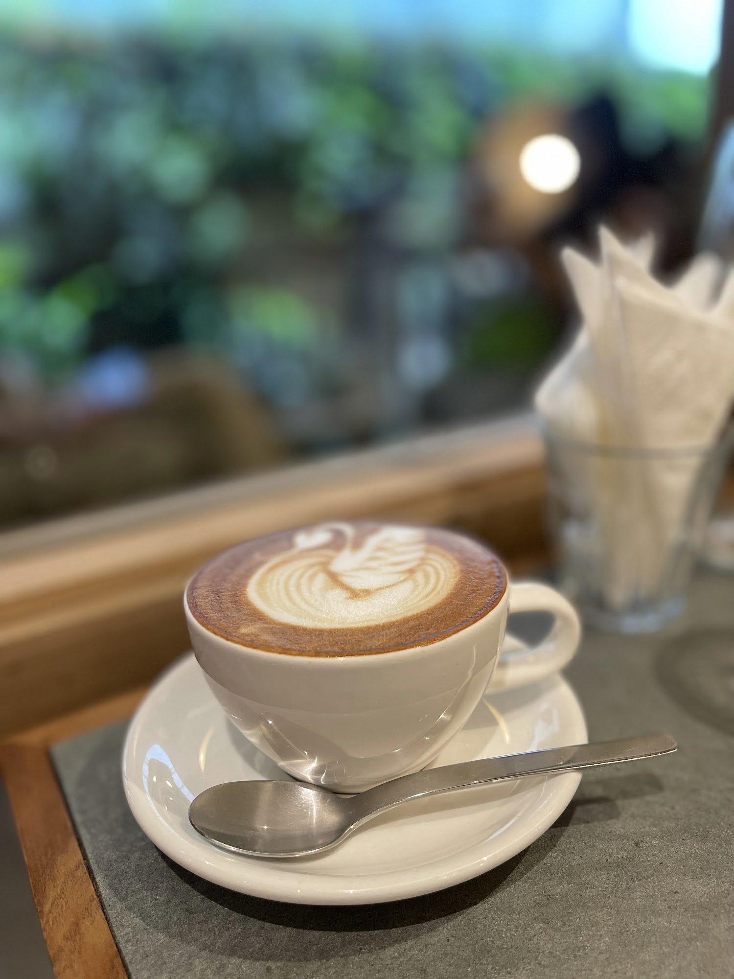 A close-up of a cappuccino in a white cup and saucer on a table with a spoon beside it, featuring latte art that resembles a swan.