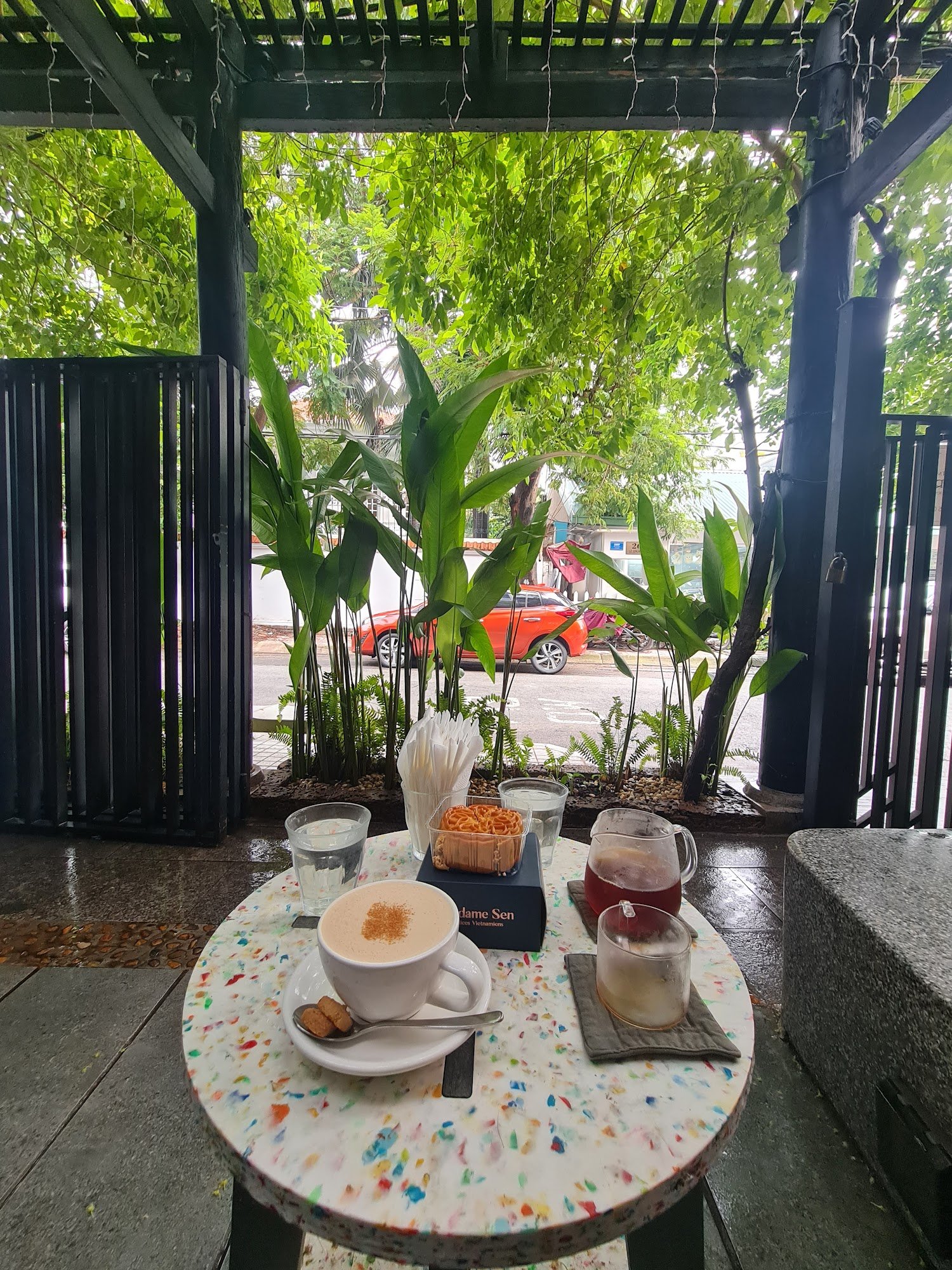 The image shows an outdoor seating area of a café or restaurant in Thao Dien, Saigon. The table is set with various beverages and snacks on a colorful