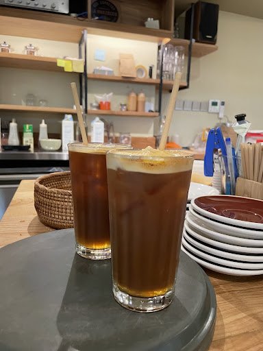 The image shows a counter in what appears to be a café or coffee shop setting with two glasses of iced coffee on the foreground, complete with straws 