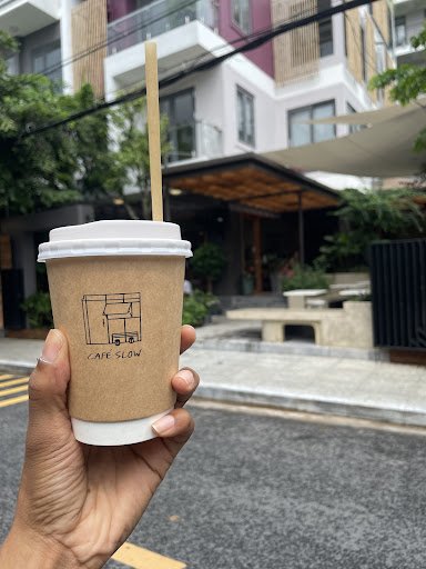 A hand is holding a takeaway coffee cup from 'Cafe Slow' in Thao Dien, Saigon. The background shows modern residential buildings with balconies and gr