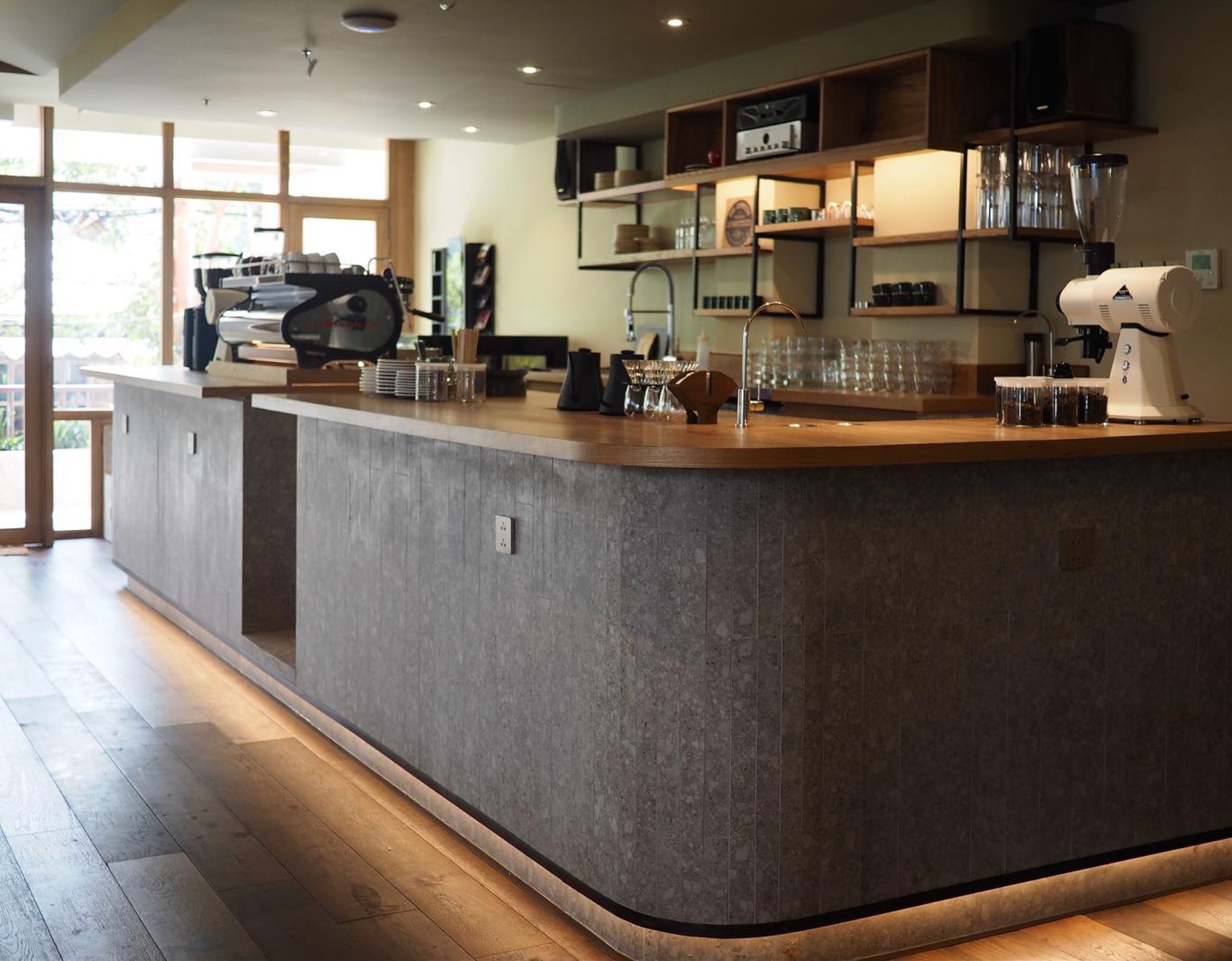 The image shows a modern coffee shop interior featuring a wooden counter and shelves stocked with various items such as cups, glasses, and small appli
