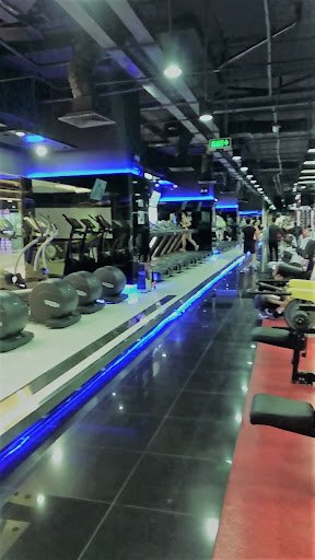The image shows an interior view of a modern gym equipped with various exercise machines and weights on display along the walls.