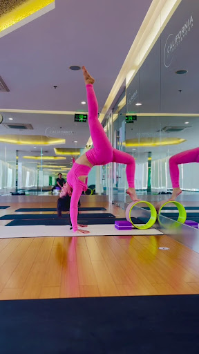 A person in a bright pink yoga outfit is performing an advanced handstand pose on a wooden floor inside a well-lit, spacious indoor gym with mirrored
