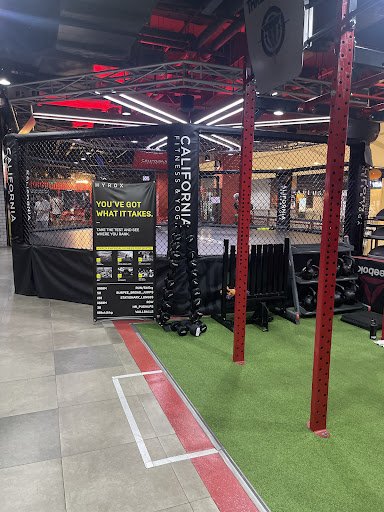 The image shows an indoor gym or fitness center featuring a boxing ring branded 'California Fitness'. The flooring is red and green, with white markin