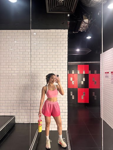 A woman is standing in a modern fitness studio wearing pink workout attire and holding a yellow water bottle with red accents.