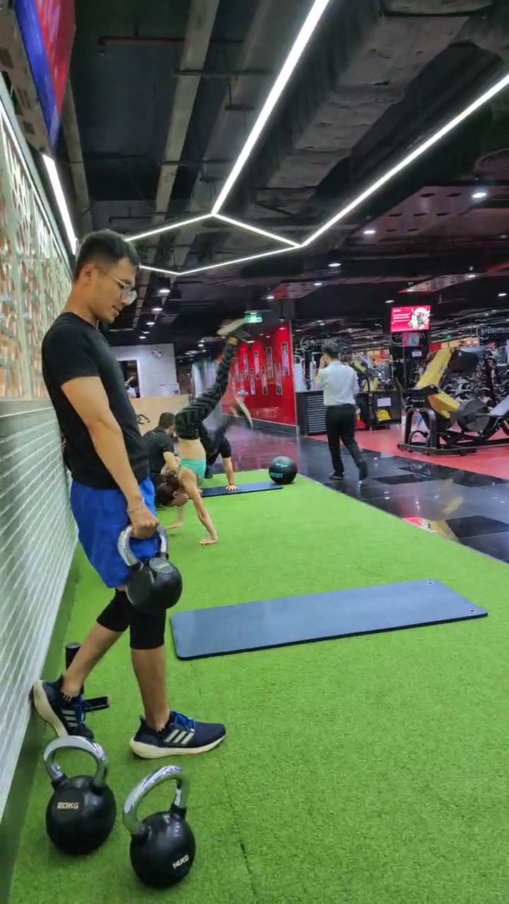 A modern gym interior in Thao Dien, Saigon, featuring green artificial turf flooring. In the foreground, a man in a black t-shirt, blue shorts, and bl