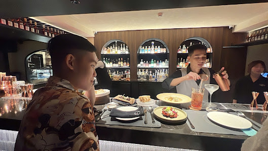 The image shows an indoor setting of a bar or restaurant where patrons are seated at the counter and being served by bartenders.