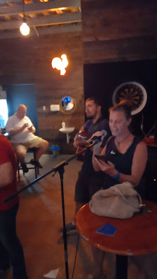 A group of people are gathered in a dimly lit room where one person is playing an acoustic guitar while another holds a microphone, possibly singing o