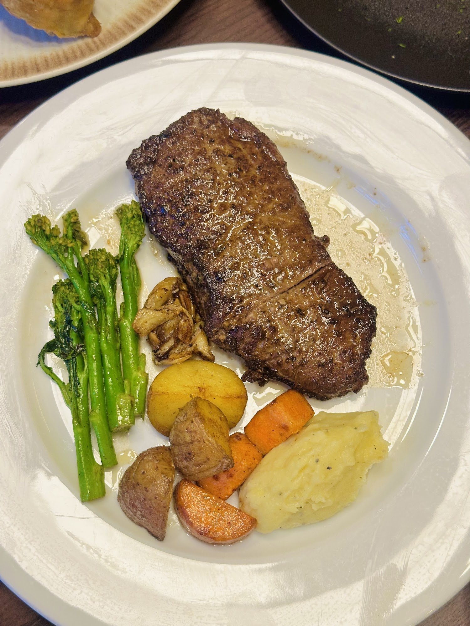 A plated meal consisting of a medium-rare steak accompanied by roasted vegetables including broccoli and potatoes, as well as what appears to be mashe