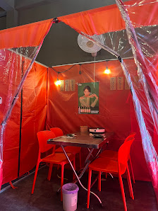 An indoor venue with orange plastic tents forming a canopy structure. Features a metal table with two red chairs, a wall-mounted fan, a poster with a 