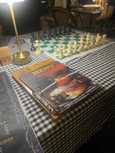 The image shows a chess game in progress on a table covered with a checkered cloth at Thao Dien, Saigon. A menu is prominently displayed alongside the