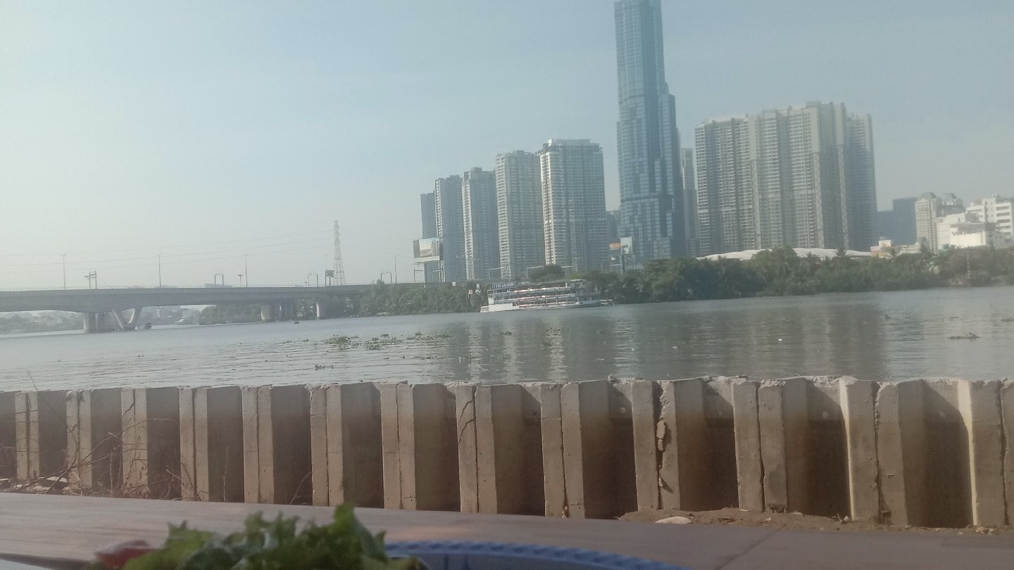 The image shows a view of the Saigon River from Thao Dien, featuring high-rise buildings in the background and concrete barriers along the riverbank.