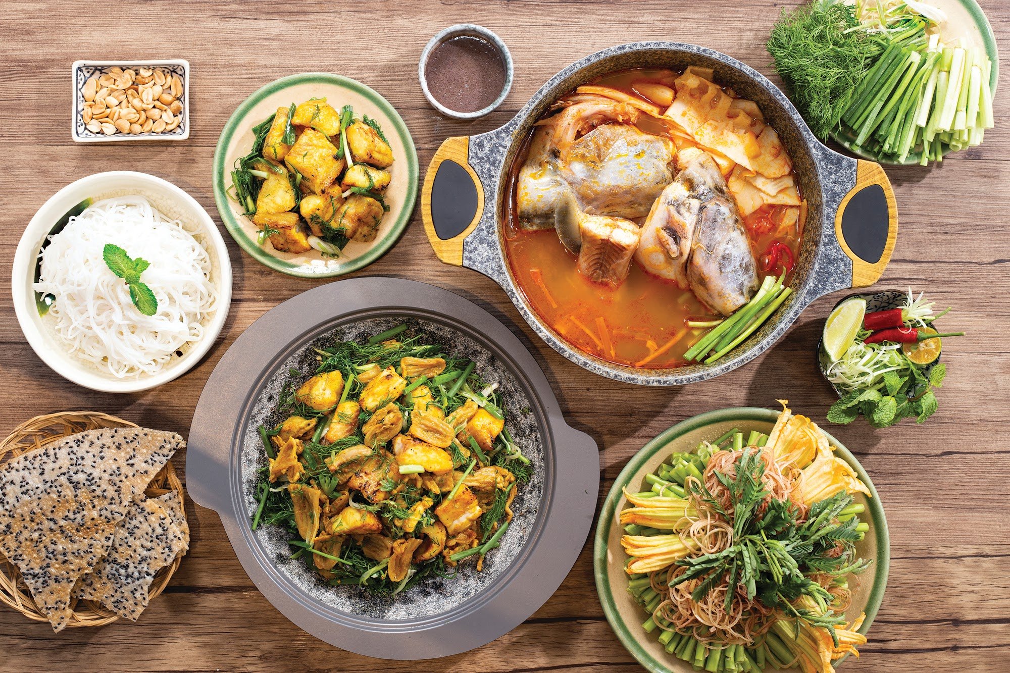 A variety of Vietnamese dishes are displayed on a wooden table including noodle soup, stir-fried tofu with herbs, fish cakes in curry sauce, and vario