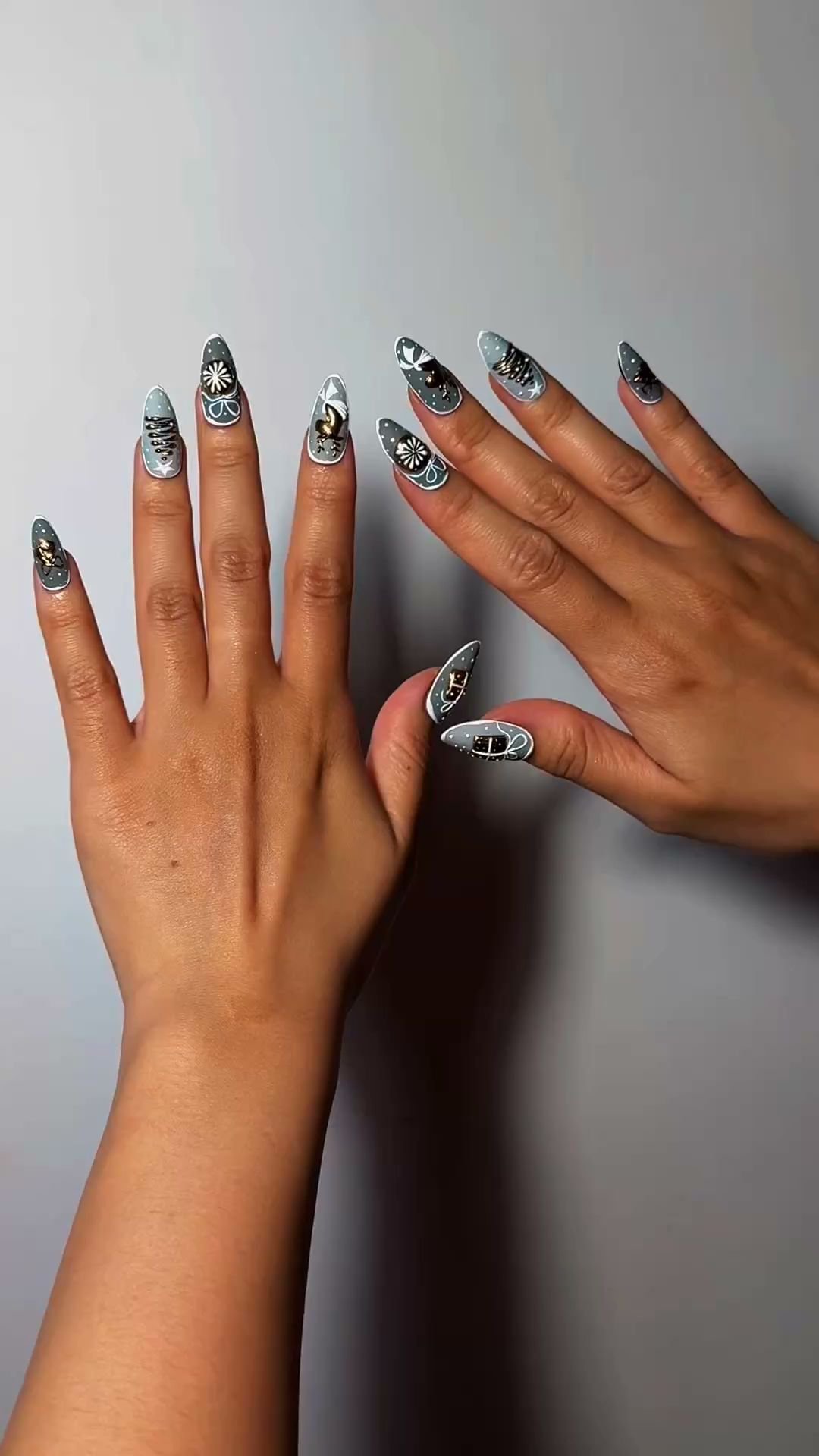 Two hands with intricately designed nail art featuring floral and animal motifs on a light blue base, displayed against a plain gray background. The n