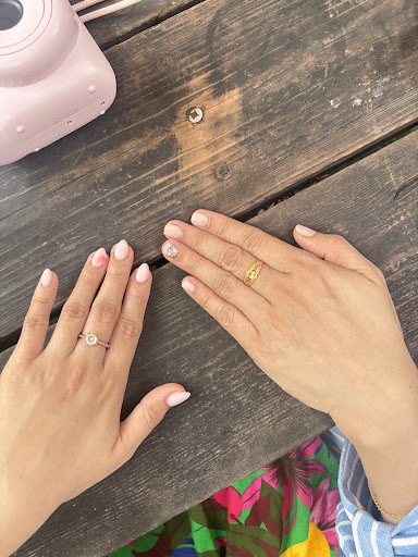 A close-up of a person's hands adorned with rings on their fingers resting on a wooden table surface in what appears to be an indoor setting, possibly