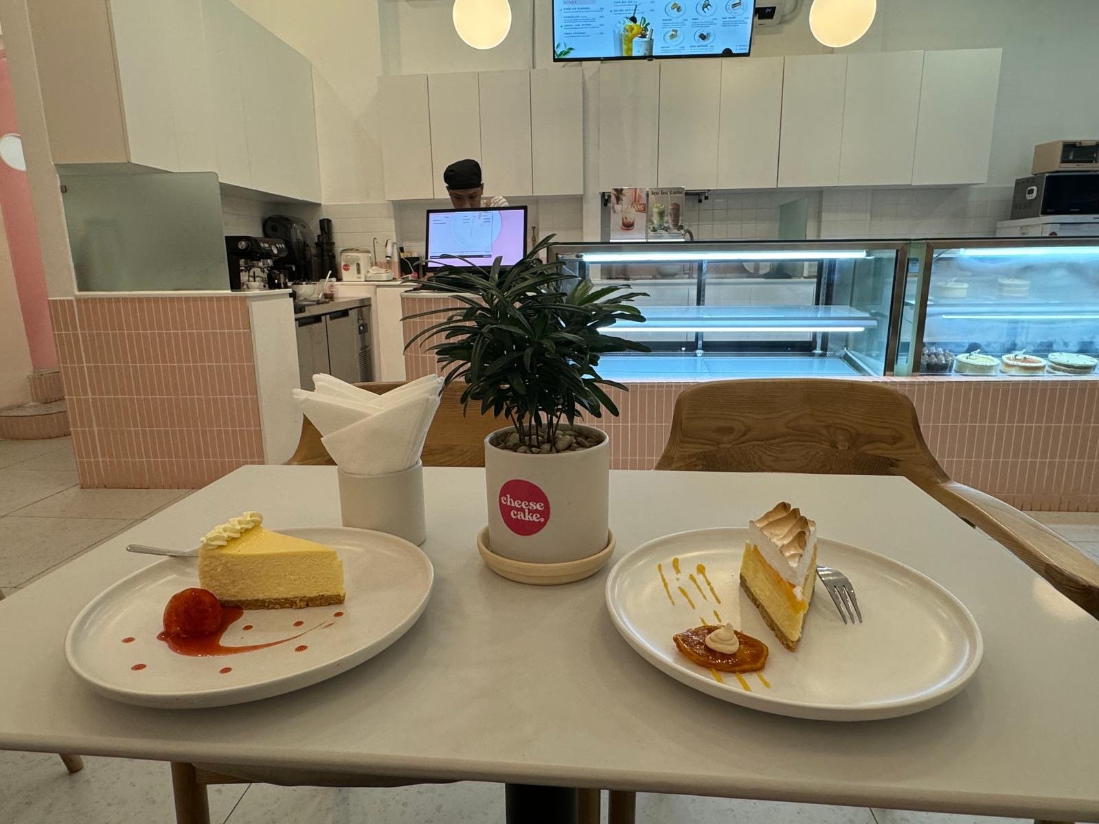 The image shows an interior view of a modern cafe or dessert shop in Thao Dien, Saigon. The focus is on two plates with slices of cheesecake served on
