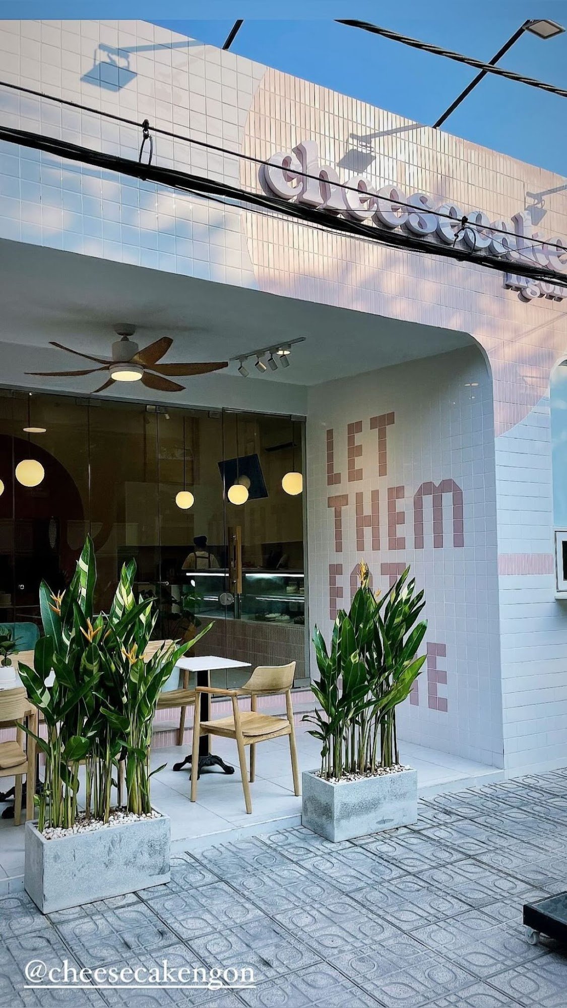 The image shows an exterior view of a cafe named 'Cheese Cake Envy' with the slogan 'LET THEM EAT CAKE' on its facade. The establishment has large win