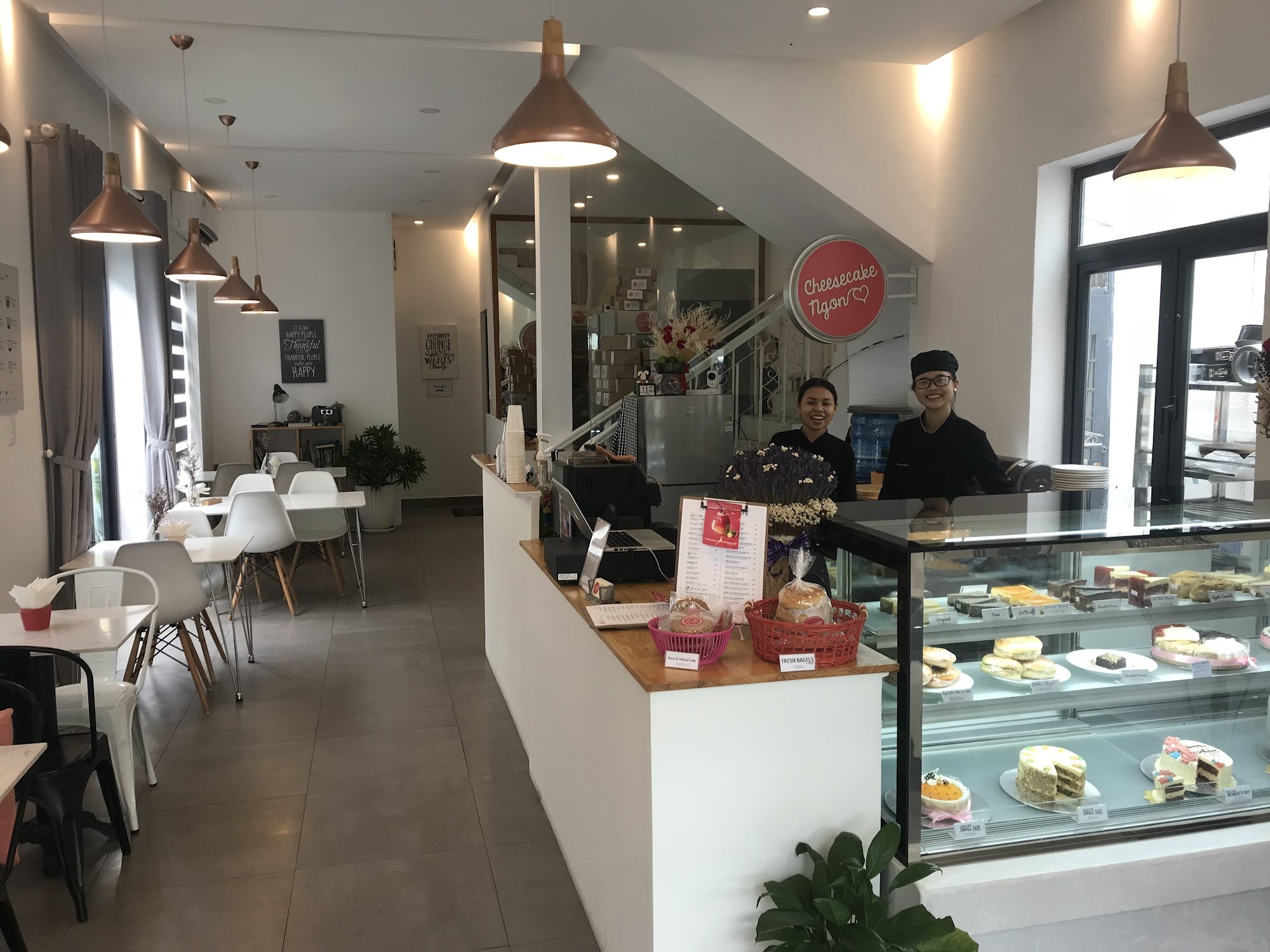 The image shows an interior view of a modern cafe or bakery named 'Cheesecake Ngon'. The establishment has a clean and contemporary design with white 